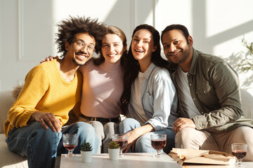Fototapeta premium Happy smiling diverse multinational friends hugging looking at camera sitting on sofa in living room