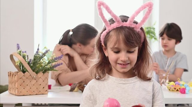 The girl with rabbit ears closes her eyes with painted Easter eggs, plays in prying eyes and smiles at the background of a large family at home