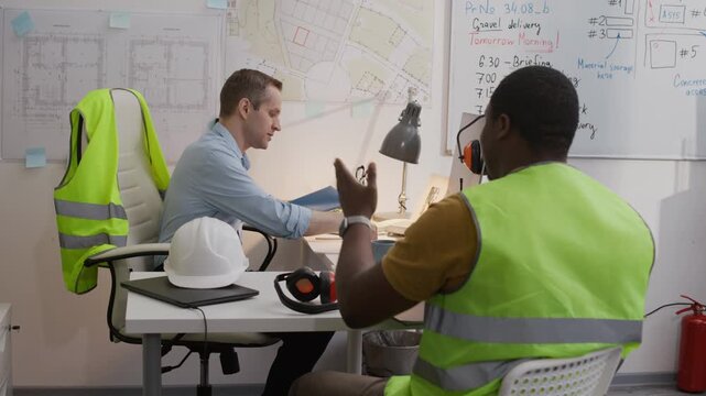 Medium long shot of black construction worker in safety vest giving building plan to Caucasian engineer sitting at office desk with blueprints on wall in background