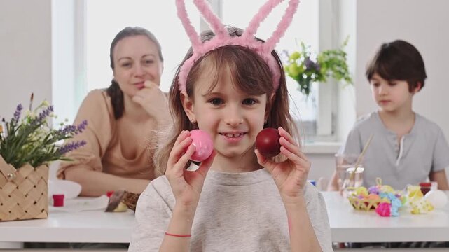 The girl with rabbit ears closes her eyes with painted Easter eggs, plays in prying eyes and smiles at the background of a large family at home
