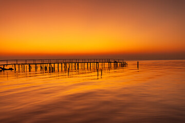Obraz premium Golden Hour Silhouette of a Wooden Pier over Calm Ocean Water,A serene and minimalist landscape featuring a long wooden pier silhouetted against a vibrant orange sunset. The calm sea perfectly.