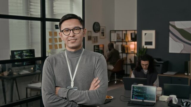 Waist-up portrait of Caucasian gen Z male software developer wearing glasses and badge folding arms and posing for camera with smile in modern office