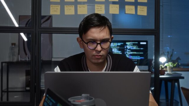Concentrated Caucasian dark-haired young IT specialist wearing glasses creating software application while sitting in front of laptop screen in office at nighttime