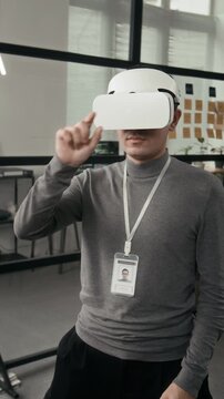 Vertical view of young tech specialist wearing grey turtleneck, badge and VR headset creating interactive environment in office