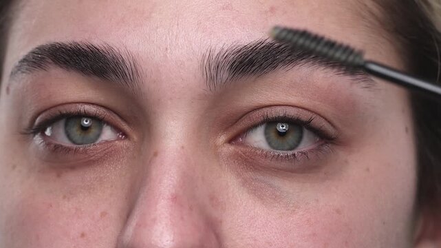 4K video closeup portrait of young woman carefully brushing and shaping her eyebrows with precision, no makeup and natural expression, studio shot on white background