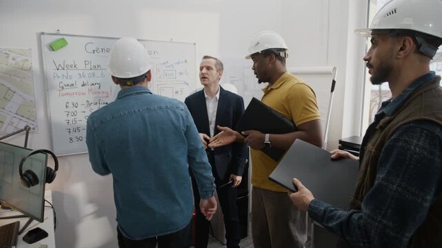 Long shot of team of four male construction engineers meeting at office, standing by whiteboard, planning and discussing work