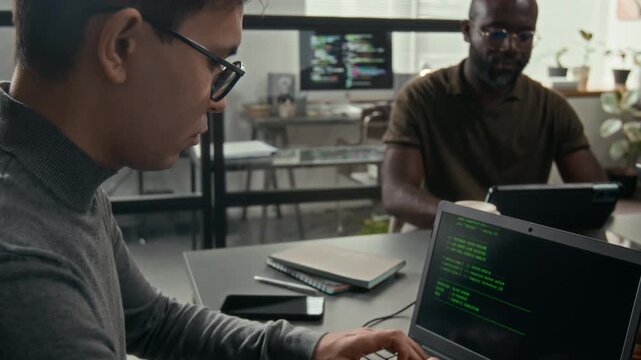 Tilt up view of young male web developer wearing glasses programming on laptop and listening to male coworkers speaking in background in IT office