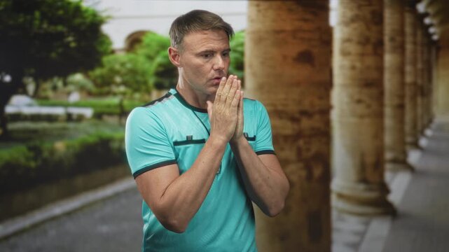 Man referee with whistle presses palms together near mouth in building corridor with stone columns; concern prayer.