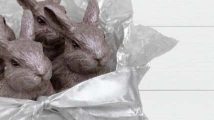 Three fluffy rabbits nestled inside a silver gift bag with a large bow, highlighting soft fur textures and metallic wrapping in bright daylight.