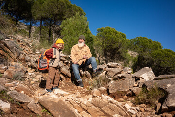 Senior grandfather resting on rocky mountain trail with young grandson during hiking adventure, family bonding, active lifestyle, outdoor travel, intergenerational connection 