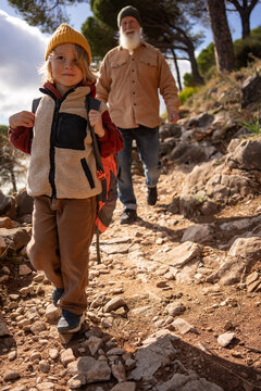Confident young boy hiking downhill on rocky mountain trail with backpack, followed by smiling senior grandfather, family adventure, outdoor bonding, active lifestyle and generational connection