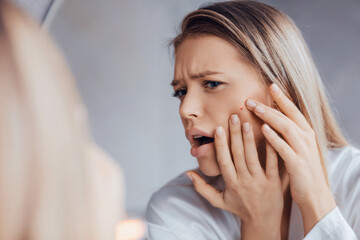 A woman stands in front of a mirror looking closely at her face. She touches a blemish on her cheek with a worried expression. Soft light fills the room. © Prostock-studio