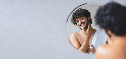 A man stands in front of a round mirror in a bathroom. He smiles as he touches his face. The setting has soft morning light and a clean, simple design. He is wrapped in a towel. © Prostock-studio