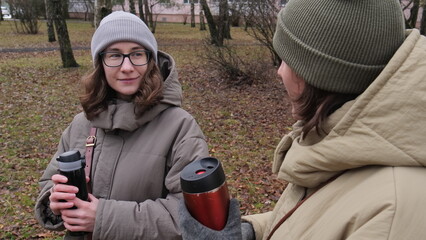 Fototapeta premium Two young women friends spending time together outdoors, sharing a warm drink from thermal mugs while having a private conversation in an autumn park setting