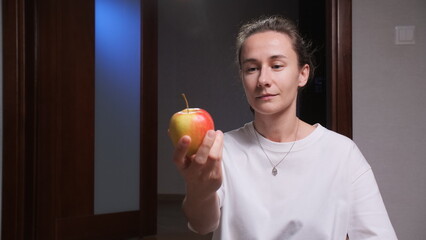 Woman stands indoors holding a fresh apple, contemplating a healthy snack and mindful choice for balanced diet and well being in a simple, natural light domestic setting © Aliaksandr