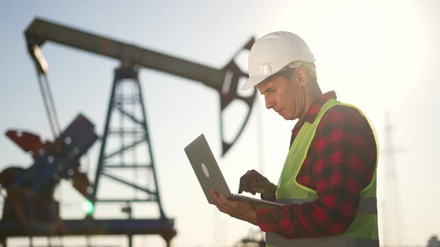 Worker checks laptop on oil field while engineer monitors pumpjack and rig operation under bright sun wearing hard hat and safety vest showing energy industry workflow and worker laptop coordination
