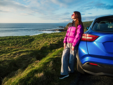 Young woman in colorful jacket is standing by a blue rented car and looking at stunning nature scenery with rough ocean coast at sunset. Mullaghmore, county Sligo. Travel and tourism in Ireland.