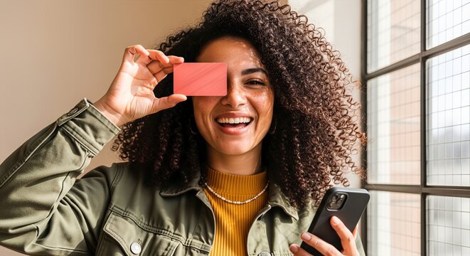 Happy young woman with curly hair holding a blank pink credit card over one eye while using her smartphone indoors.