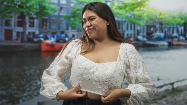Woman adjusting blouse with hands on hem on street canal near boats and railing; confidence relaxed.