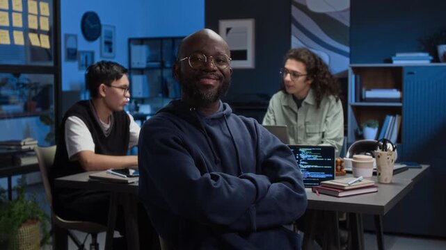 Portrait of African American millennial web developer wearing hoodie smiling for camera with crossed arms while working at office desk with two Caucasian colleagues in background