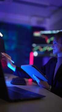 Cybersecurity analyst monitoring real-time network threat logs in dark security operations center. Digital forensic expert auditing malware code syntax via tablet interface.