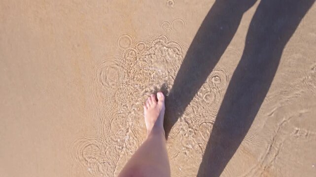 A woman's bare feet walking along the beach, in shallow water, shadow from the sun, top view