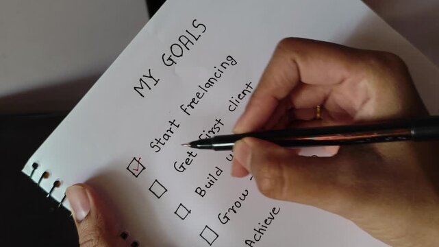 Close up video of a person writing and completing freelancer goals checklist in a notebook, including start freelancing, get first client, build website, and grow income, with achieve freedom circled.