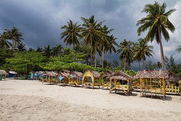 Traditional wooden beach huts on sandy shore with tall palm trees and dramatic cloudy sky in Bintan Indonesia.