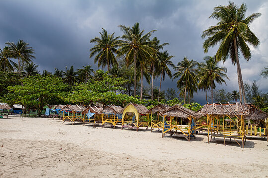 Traditional wooden beach huts on sandy shore with tall palm trees and dramatic cloudy sky in Bintan Indonesia.