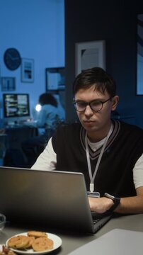 Vertical view of fatigued gen Z QA specialist wearing glasses testing application on laptop in dimly lit IT office with cookies on plate on his desk