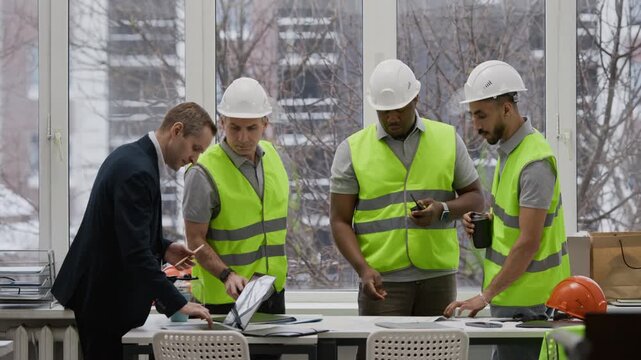 Medium long shot of multiethnic construction team in vests and hardhats reviewing renovation plan with male engineer in suit by table in office