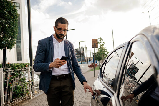 Urban professional using smartphone to request or access ride hailing transportation beside a parked car in a modern city environment, representing app mobility and daily commuting.