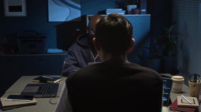 Caucasian gen Z programmer chatting with positive Black millennial QA specialist while working on laptops sitting at grey tables opposite each other