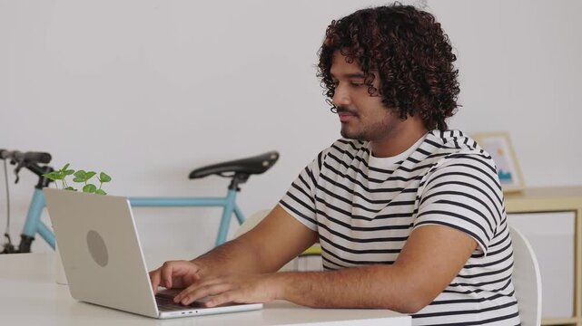 Young adult Indian man working on laptop at home office desk. Remote professional using technology in modern interior. Business concept.