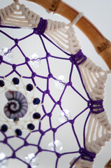 Handmade macrame dream catcher with bamboo hoop, cotton rope and violet cord, decorated with crystal beads and spiral shell. Boho wall hanging on neutral background.
