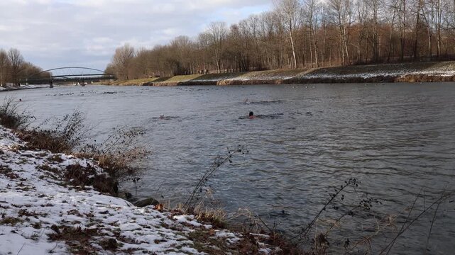 Cold season river marathon in urban park swimmers spread across wide channel battling icy current while pedestrians pause along frosted embankment and steel arch bridge rises in background