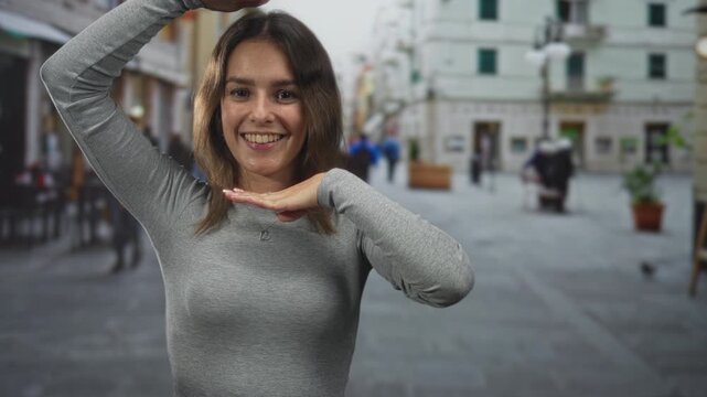 Woman smiling and posing with hands framing her face and palms up on a cobblestone street plaza in the city; joy travel.