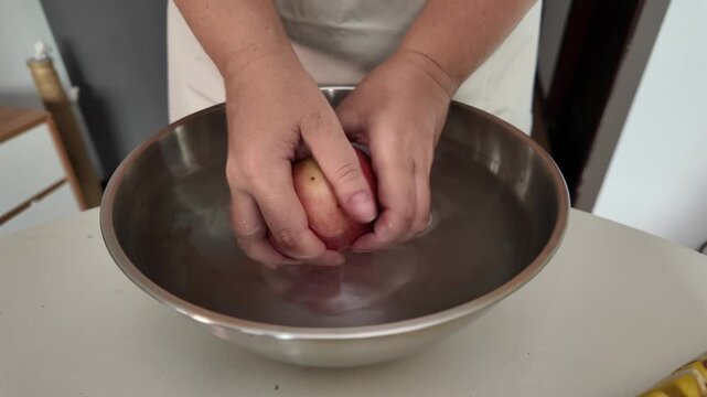 Close-up of a woman's hands in an apron, standing in front of a table with fruit and washing red apples