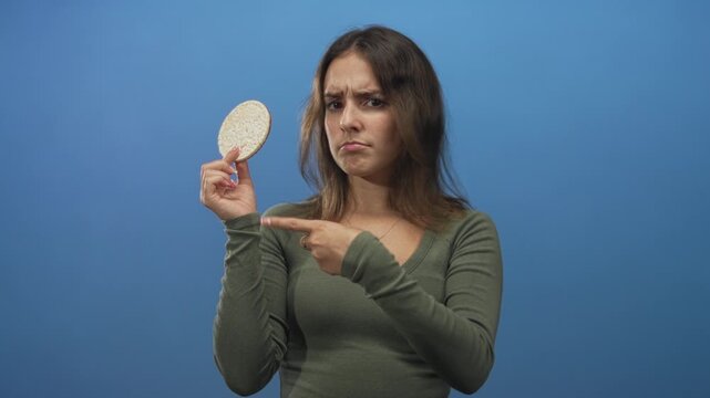 Woman holding a round rice cake with one hand and pointing finger at it with the other in blue studio; skepticism doubt.