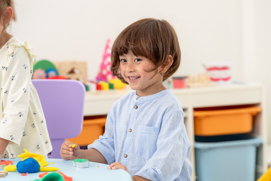 Toddler boy smiles during preschool playroom activity, holding playdough piece, waiting turn, learning patience, single mother supervising nearby, family bonding activity without screen, indoor joy