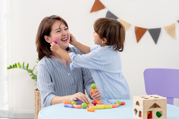 Naklejka premium Asian single mother smiles during pretend makeup game, preschool boy touches face using colorful toy, teaching gentle care, patience, respect, parent child interaction, cozy indoor family playtime.