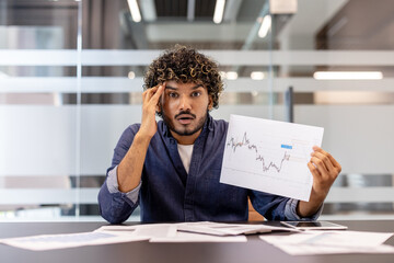 Photo of shocked young Indian office worker sitting at desk. pointing at camera at graphs and holding head