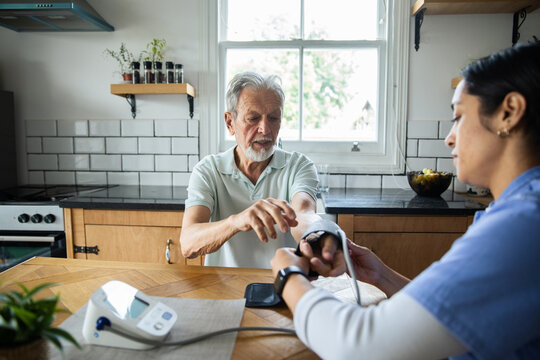 Home nurse checking senior man's blood pressure in kitchen