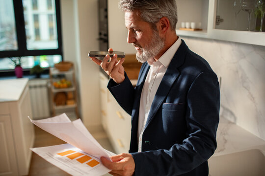 Mature businessman using voice assistant in home kitchen while reviewing financial reports