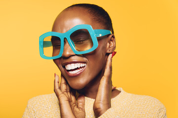 Happy African woman with dark skin wears large blue glasses and a beige sweater, smiling with eyes...