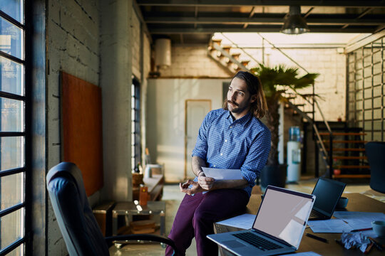 Thoughtful young man holding documents in modern loft office