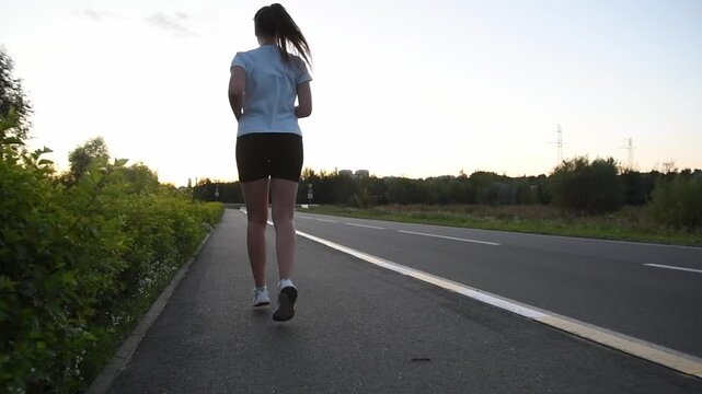 Young woman running away from camera on sidewalk at sunset filmed from low ground level in slow motion. Back tracking shot emphasizes powerful strides and dynamic urban fitness atmosphere.