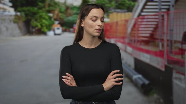 Woman with arms crossed and eyes closed on a city street by red construction fence; indifference defiance.