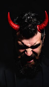 Man with dark beard and red devil horns grimacing with teeth bared against black background