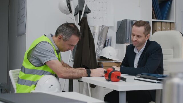 Medium long shot of middle-aged male construction worker getting payment from manager while sitting at table in small office